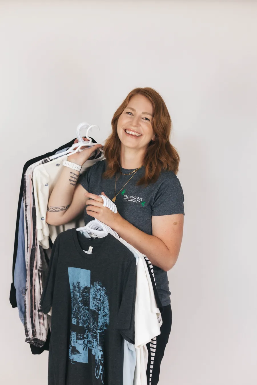 Kristen laughing in a KAS KONCEPTS t-shirt, holding an armful of clothes on hangers