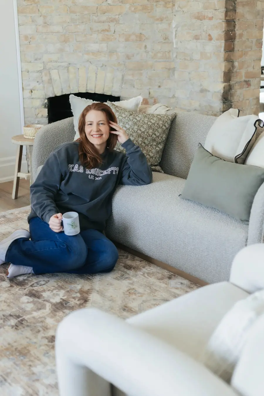 Kristen sitting on the floor against a sofa, holding a mug and wearing a KAS KONCEPTS sweatshirt