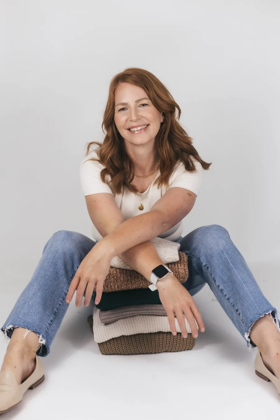 Kristen, founder of KAS KONCEPTS, smiling while sitting with her arms crossed over a neat stack of folded sweaters